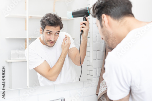 Young handsome man standing in front of the mirror in the bathroom and holding hair dryer. Morning routine and making hair style concept