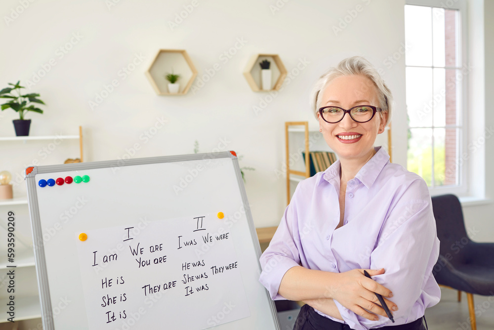 English teacher in school classroom. Happy beautiful elegant middle aged woman in shirt and eyeglasses standing with her arms crossed by white board with grammar rules, looking at camera and smiling English teacher in school classroom. Happy beautiful elegant middle aged woman in shirt and eyeglasses standing with her arms crossed by white board with grammar rules, looking at camera and smiling