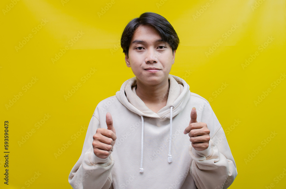 handsome attractive Asian man smiling happily and giving thumbs up as compliment, isolated over yellow background. man in beige hoodie giving approval and compliment by gesturing thumbs up. handsome attractive Asian man smiling happily and giving thumbs up as compliment, isolated over yellow background. man in beige hoodie giving approval and compliment by gesturing thumbs up.