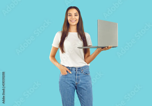 Portrait of smiling young woman who holding modern laptop pc computer that she uses for work, study and leisure. Woman in t-shirt and jeans posing with laptop isolated on light blue background.