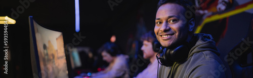 Positive indian gamer with headphones looking at camera in cyber club, banner.