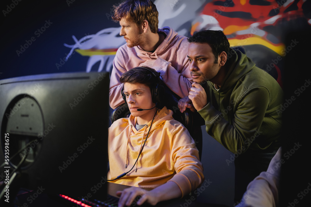 Fototapeta premium Focused indian man standing near friend playing video game on computer in gaming club.