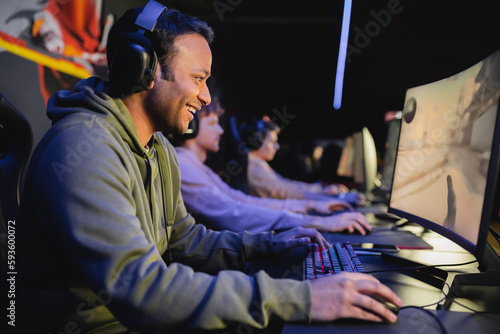 Side view of smiling indian man in headphones playing video game on computer with blurred team in cyber club.