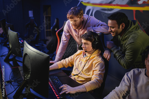 Multiethnic friends standing near gamer in headphones playing video game on computer in cyber club.