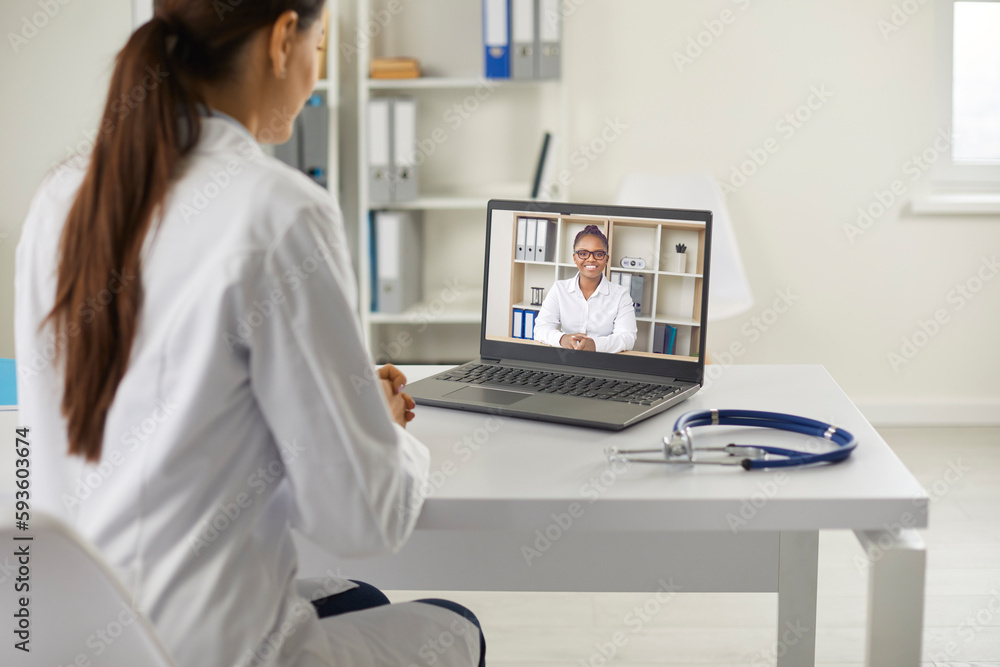 Doctor using laptop and giving online video consultation to patient. Female physician sitting at white table with notebook computer and giving medical advice to young woman via videocall Doctor using laptop and giving online video consultation to patient. Female physician sitting at white table with notebook computer and giving medical advice to young woman via videocall