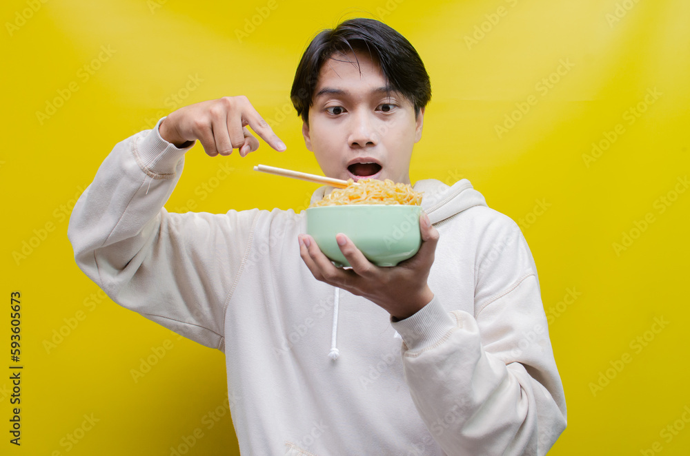 Closeup an Asian man in a beige hoodie eats instant noodles using chopsticks and a bowl, isolated over a yellow background. Closeup an Asian man in a beige hoodie eats instant noodles using chopsticks and a bowl, isolated over a yellow background.