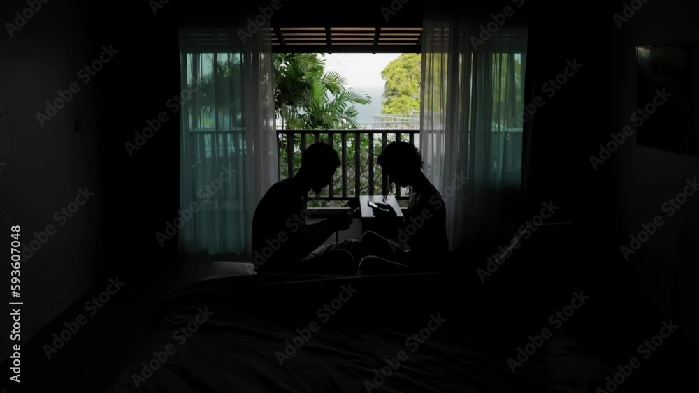 Man and woman are using smartphones sitting together in hotel room near open balcony. Silhouettes of couple surf internet and read news in social network not talking to each other. Virtual addiction Man and woman are using smartphones sitting together in hotel room near open balcony. Silhouettes of couple surf internet and read news in social network not talking to each other. Virtual addiction