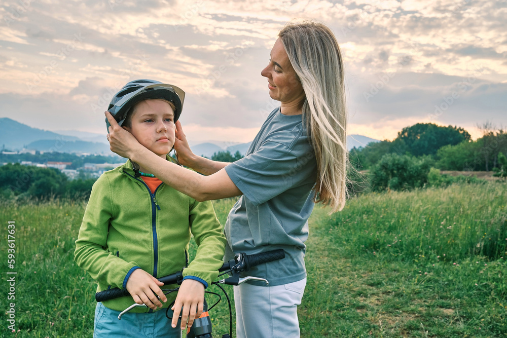 Happy Family outdoors, Mother teaching son to ride bicycle. Happy cute boy in helmet learn to riding a bike in park on green meadow in summer day at sunset time. weekend.
