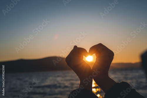Women fingers making a heart shape on the seashore at sunset. Hand heart shape silhouette with sunshine