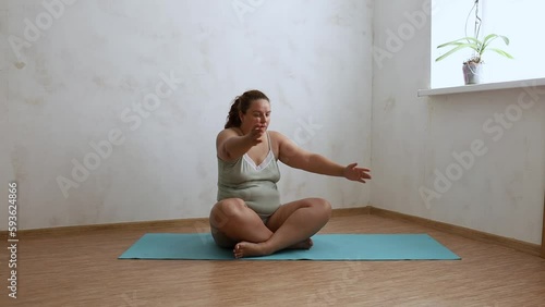 Overweight woman sitting on gym mat in empty room, take deep breaths squeezing and unclenching body and waving arms to sides. Home morning workout, sport, exercise technique, weight loss. photo