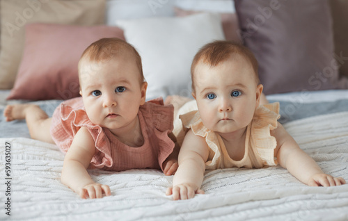 Close-up portrait of two sweet baby sisters 6 month in cotton dresses lying on comfortable bed at home. Full body fraternal twins.	
