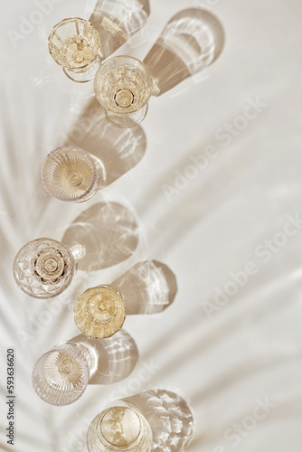 Summer party drinks flat lay, wine glasses with white sparkling wine and palm leaf shadow on light table. Minimal pattern with beautiful wine glasses, above view still life, beige golden neutral color