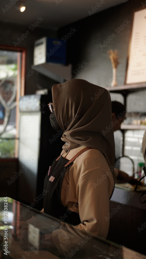 female barista wearing hijab waiting for customers behind coffee shop bar 