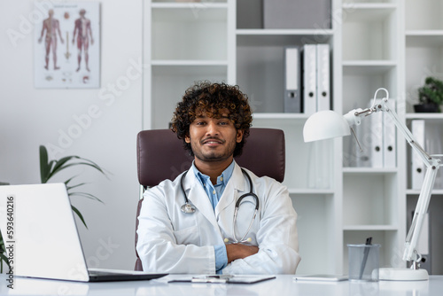 Portrait of a successful Hindu doctor inside the office of a modern clinic, sitting at the workplace at the table smiling and looking at the camera with his arms crossed, a man in a white coat.