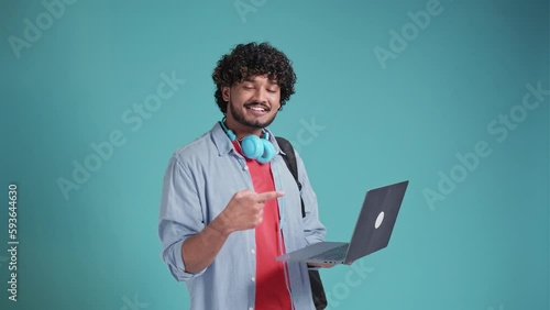Handsome Indian man student on a blue background, holding a laptop with and pointing at him with a hand gesture.