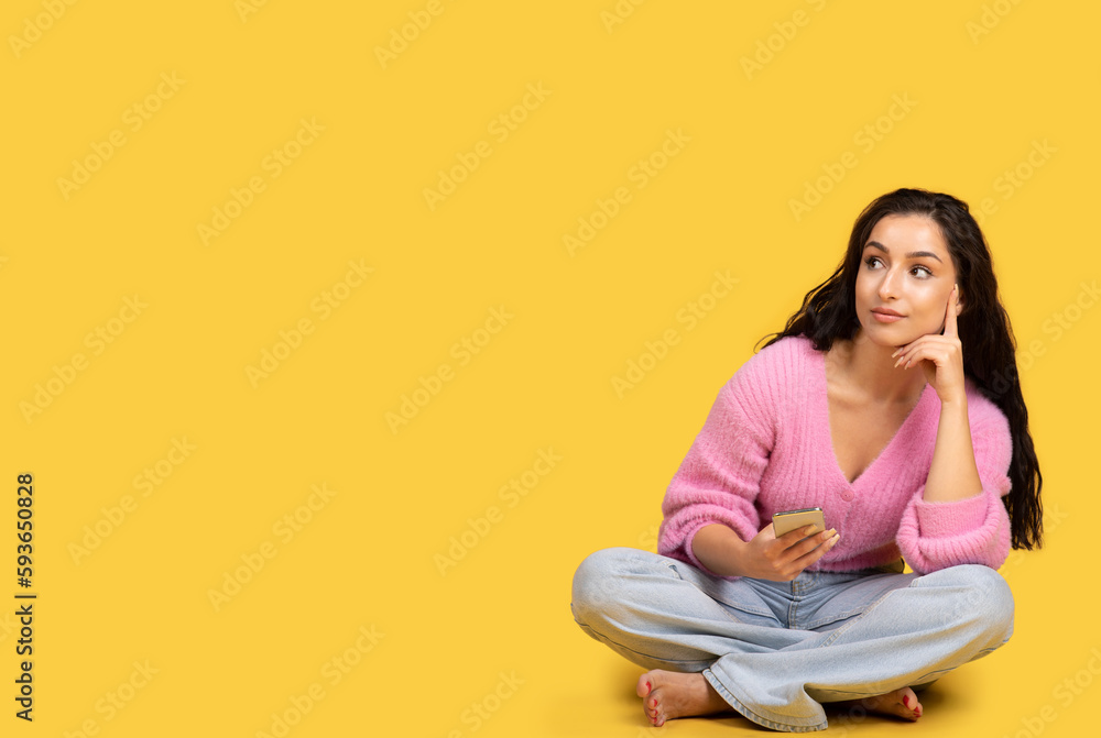 Thoughtful millennial arab barefoot lady with smartphone sits on floor and looks at copy space Thoughtful millennial arab barefoot lady with smartphone sits on floor and looks at copy space