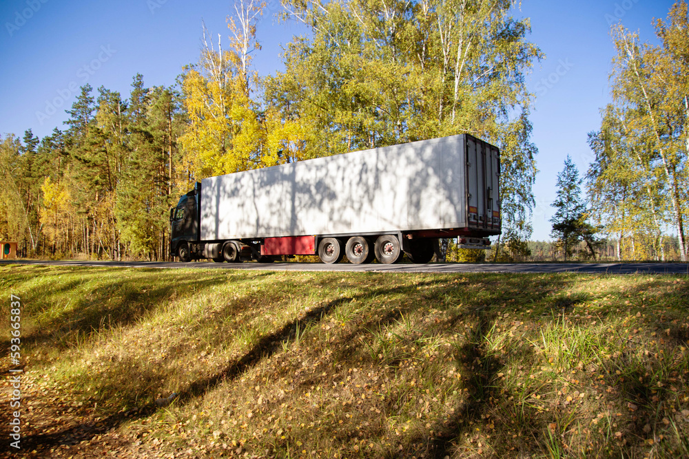 A truck with a refrigerated semi-trailer transports perishable goods against the backdrop of a forest along a country road. Copy space for text A truck with a refrigerated semi-trailer transports perishable goods against the backdrop of a forest along a country road. Copy space for text