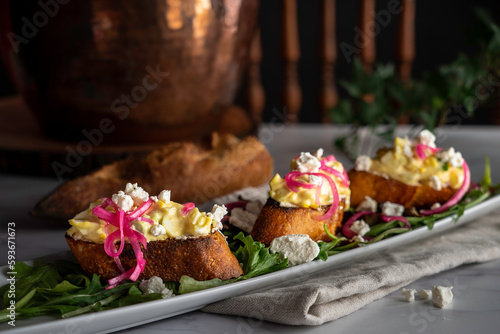 Creamy egg salad tops grilled slices of baguette on a bed of arugula. Garnished with pickled red onions and crumbled goat cheese.