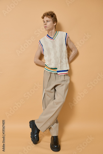 Full body length vertical shot of handsome fashion red head man model wearing stylish outfit casual knitted white shirt, beige pants and black shoes standing with his legs crossed poses on yellow wall