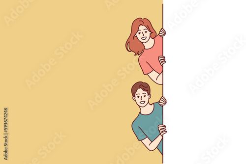 Happy man and woman with white banner peeking out recommending to pay attention to good advertisement offer. Young couple of guy and girl holding billboard for commercial or discount advertisement