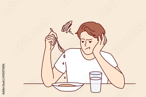 Man suffers from lack of appetite sitting at table in kitchen and does not want to eat breakfast. Depressed guy with unhappy expression melancholy looks at plate with not appetite food during diet 