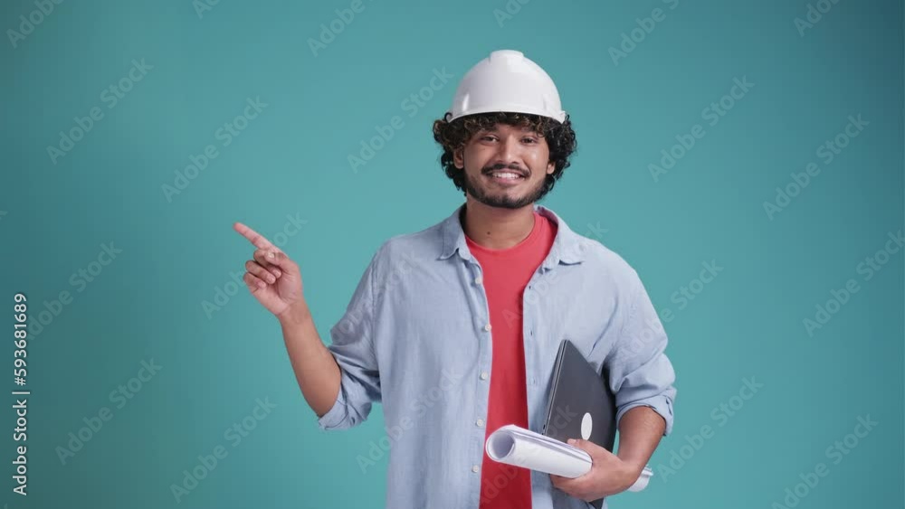 Young indian or arabic architect man with helmet and holding blueprints and laptop over isolated background pointing finger to the side and presenting a product Young indian or arabic architect man with helmet and holding blueprints and laptop over isolated background pointing finger to the side and presenting a product