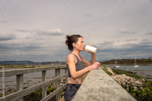 sportswoman drinking water from bottle after workout, resting on a bridge by the coast