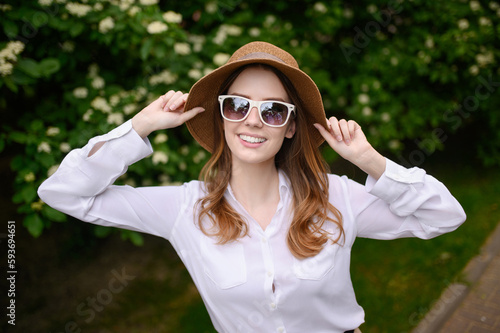 Young happy woman in summer clothes, sunglasses, straw hat posing on the background of nature, jasmine, makes a selfie portrait. Summer, technology concept