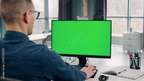 Young Man Working At Computer With Green Mock Up Screen in Office. Man Typing on Keyboard and Looking at PC Display