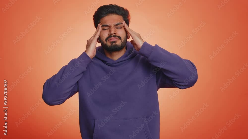 Handsome indian bearded man in hoodie rubbing temples to cure headache problem, suffering from tension, migaine, stress, grimacing in pain, high blood pressure isolated alone on orange wall background Handsome indian bearded man in hoodie rubbing temples to cure headache problem, suffering from tension, migaine, stress, grimacing in pain, high blood pressure isolated alone on orange wall background