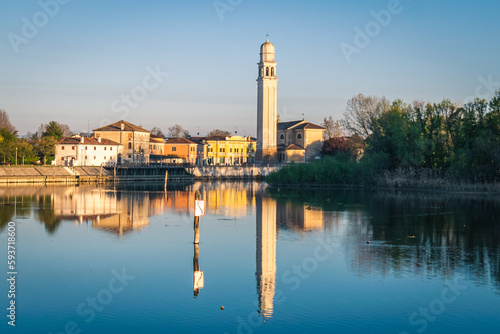 A bell tower is reflected in the water of a lake with a row of buildings in the background and a bridge in the foreground, Sile, Italy