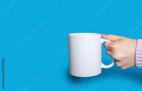 A hand holds a white cup with a hot drink on a blue background. The hand of a woman dressed in a sweater holds a large ceramic cup on a bright blue background. Free space for text