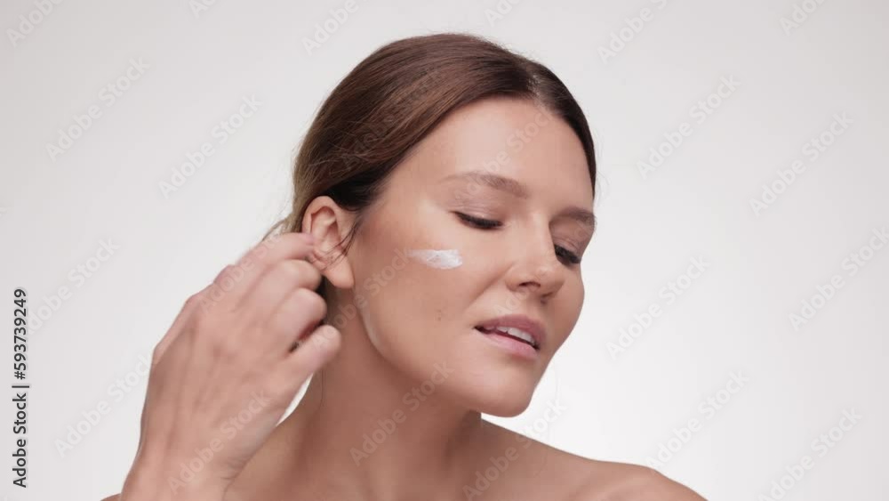 Woman doing beauty procedures on white studio background. Lady cream applying smear of pampering cream on her cheek and smiling to the camera. Skin care concept. Cosmetic Concept. Woman doing beauty procedures on white studio background. Lady cream applying smear of pampering cream on her cheek and smiling to the camera. Skin care concept. Cosmetic Concept.