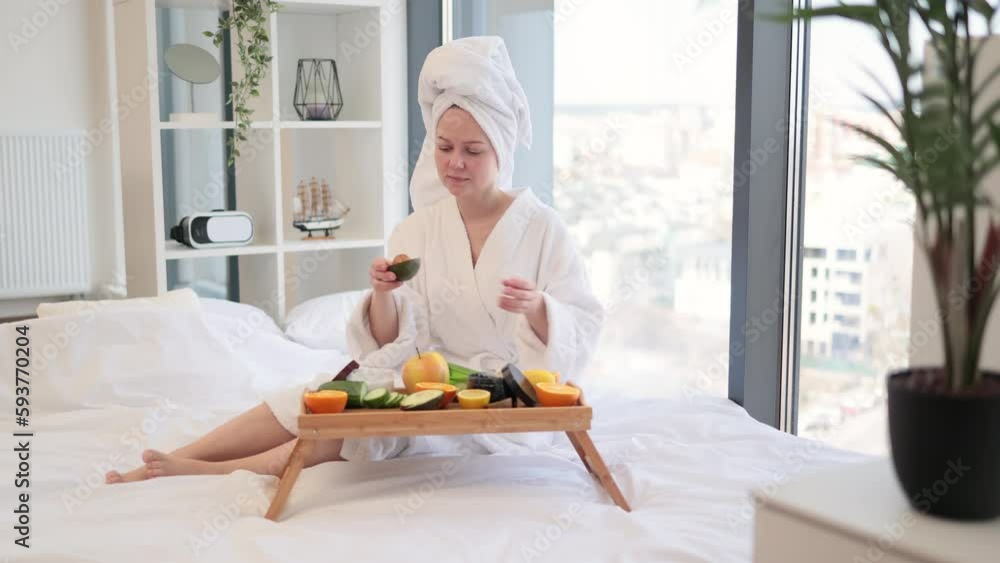 Attractive woman wearing white robe and towel on head holding avocado half and scanning cosmetic bottle while relaxing in spacious bedroom. Charming female enjoying daily skincare routine at home. Attractive woman wearing white robe and towel on head holding avocado half and scanning cosmetic bottle while relaxing in spacious bedroom. Charming female enjoying daily skincare routine at home.