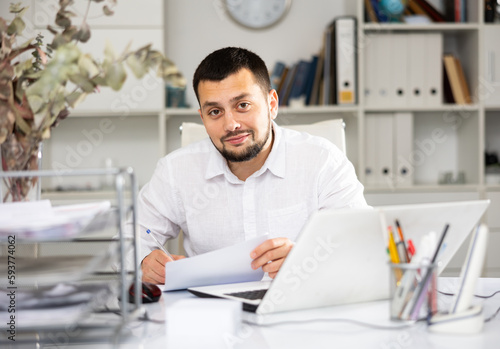 Cheerful man bookkeeper doing paperwork in his workplace in office. Smiling office manager looking at camera.