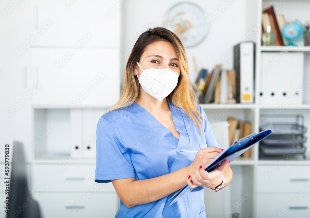 Young colombian female medical mask in uniform holding clipboard in doctor's office