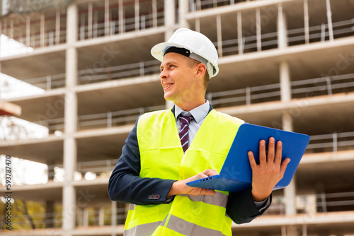European architect in workwear and white hardhat with laptop in hands standing on building site.