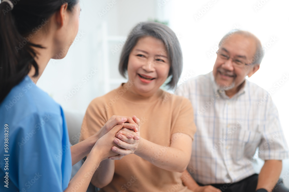 Smiling young nurse in uniform sitting with senior male and female couple at home assuring and building them in confidence about recovery in health Smiling young nurse in uniform sitting with senior male and female couple at home assuring and building them in confidence about recovery in health