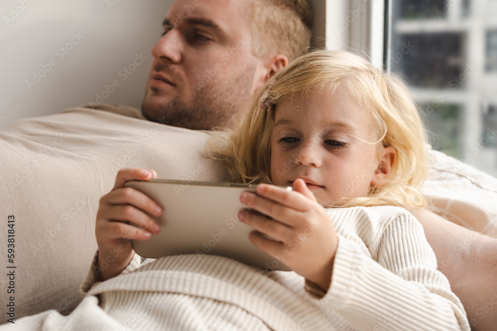 Girl and adult man lying on bed using phones at home near window. Child and father looking at gadgets, spending time together. Girl and adult man lying on bed using phones at home near window. Child and father looking at gadgets, spending time together.