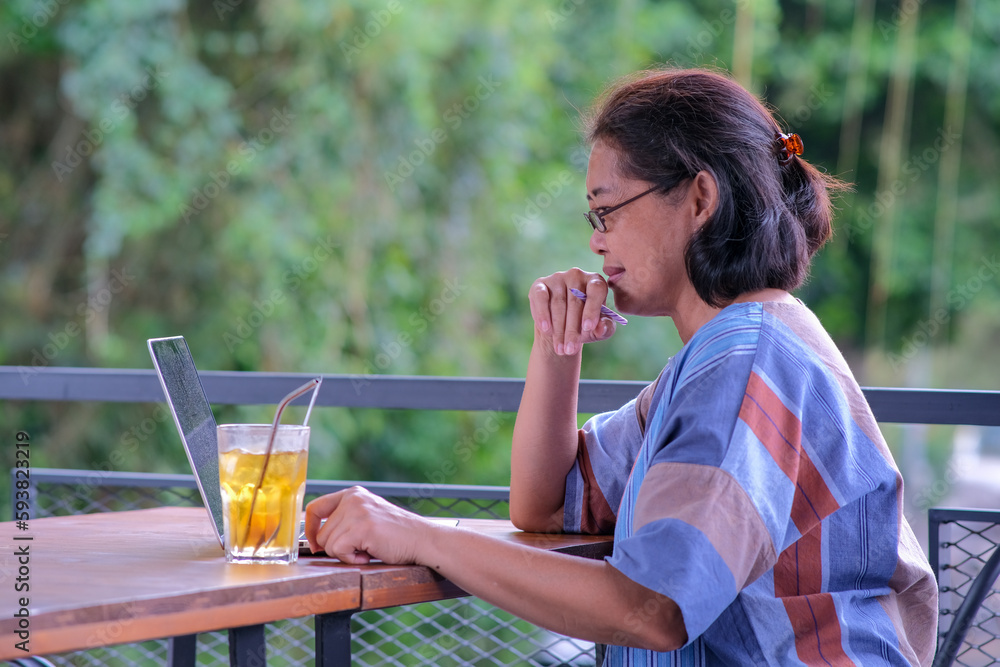 Fototapeta premium Asian woman sitting at her home balcony working on her laptop computer with a glass of lemon tea on the table