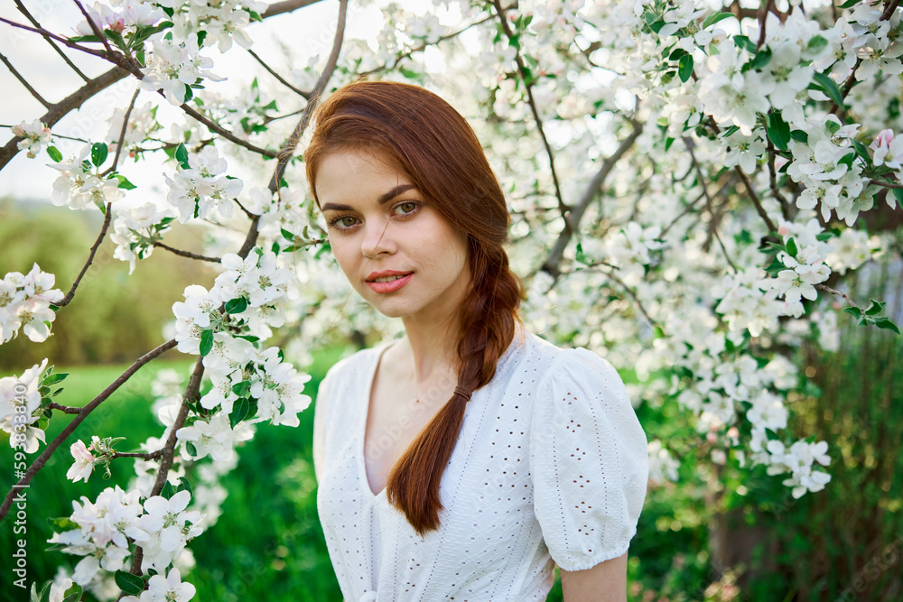 beautiful red-haired woman in a white dress smiling stands near a flowering tree beautiful red-haired woman in a white dress smiling stands near a flowering tree