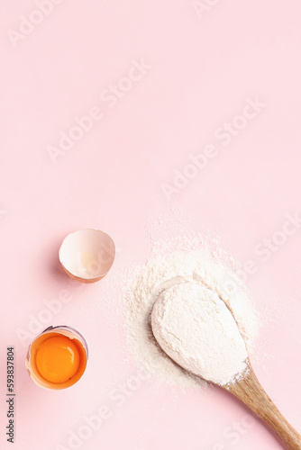 Wooden spoon with wheat flour and cracked egg on pink background