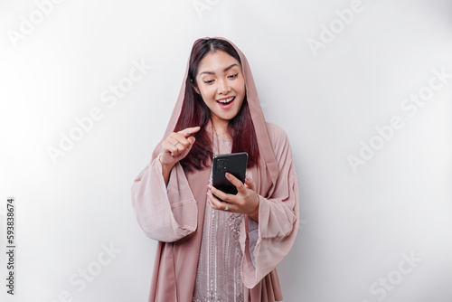A happy Asian Muslim woman wearing a headscarf, holding her phone, isolated by white background