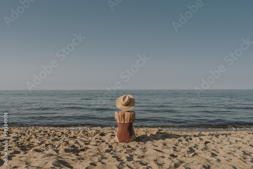 Aesthetic summer vacation fashion concept. Young tanned woman wearing a swimsuit and straw hat is sitting and relaxing on tropical beach with sand and watching at sea