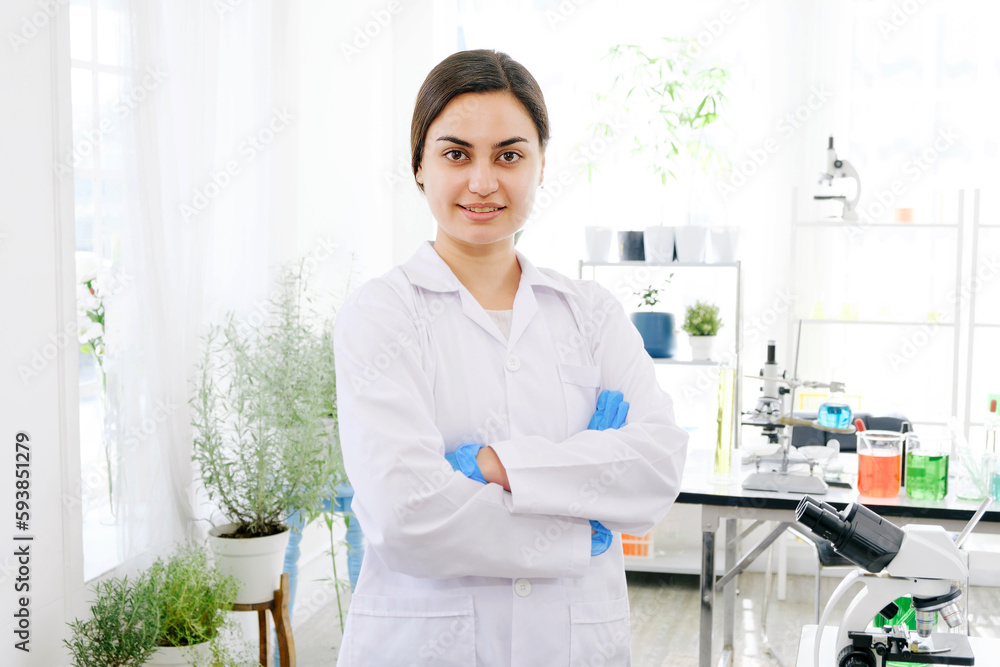 Portrait of a Middle East female scientist sitting in a laboratory and looking at the camera with the microscope, chemical liquid in beakers on the table and plants in the background. Portrait of a Middle East female scientist sitting in a laboratory and looking at the camera with the microscope, chemical liquid in beakers on the table and plants in the background.
