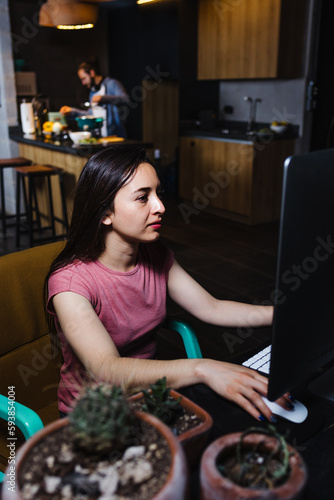 Young latin woman student working and using computer while boyfriend cooking on background at home at night in Mexico Latin America, hispanic people