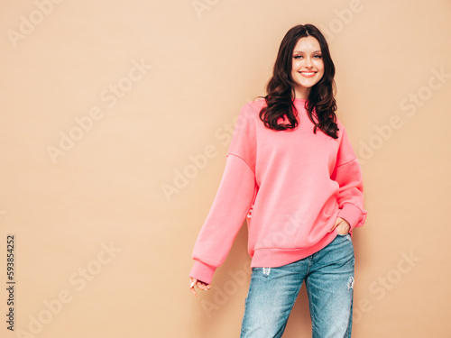 Young beautiful smiling female in trendy summer jeans and pink sweater clothes. Sexy carefree woman posing near beige wall in studio. Positive model. Cheerful and happy