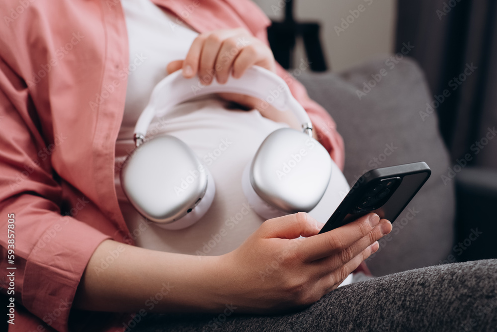 Fototapeta premium CLose up of young pregnant woman applying headphones on belly headphones enjoying relaxing music, listening to playlist from smartphone, radio. Peaceful expectant mom taking tranquility sound therapy