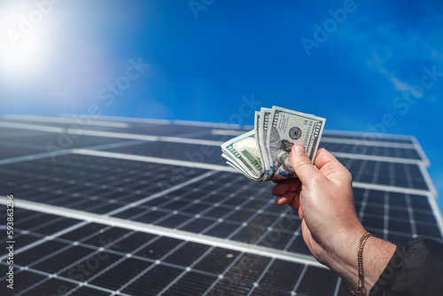 Saving money with solar energy and solar panels. Hand with dollars in front of panels.