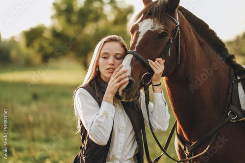 Young beautiful female jockey strokes and hugs the horse's head and prepares for the competition. Jumping training in the meadow in summer evening. © sergo321